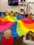 Dzieci z grupy Gwiazdeczek wraz z Panią wychowawczynią machają kolorową chustą z materiału